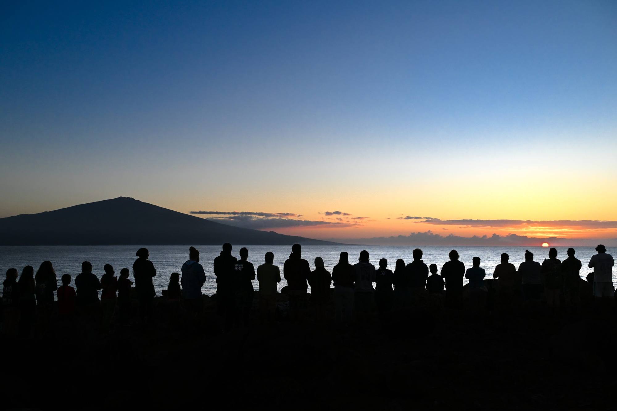 Silhouettes of people stand along a shoreline at sunrise, with a mountain and calm water in the background.