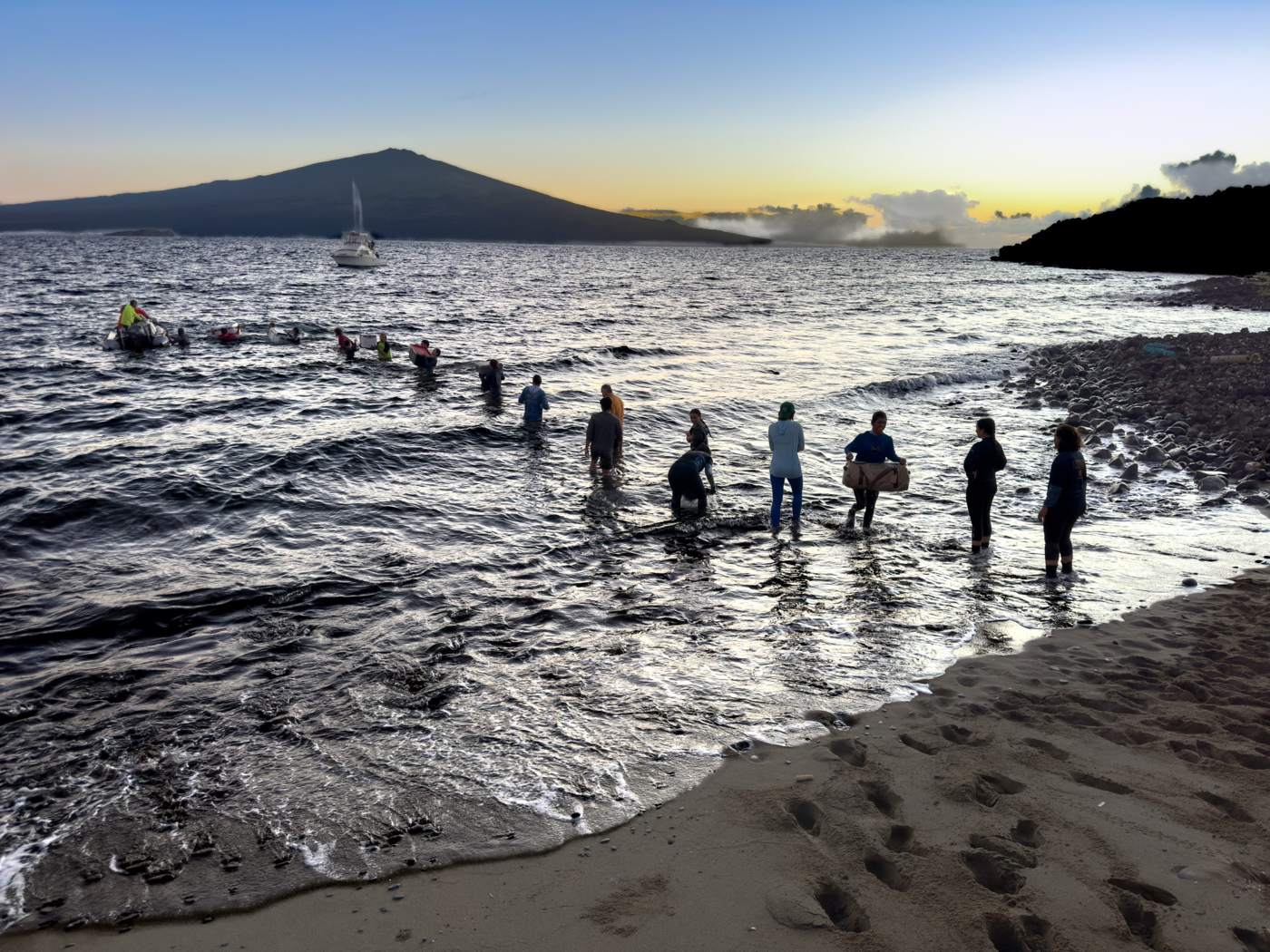 People stand in shallow water near a rocky shoreline at sunset, with a boat and a distant mountain visible in the background.