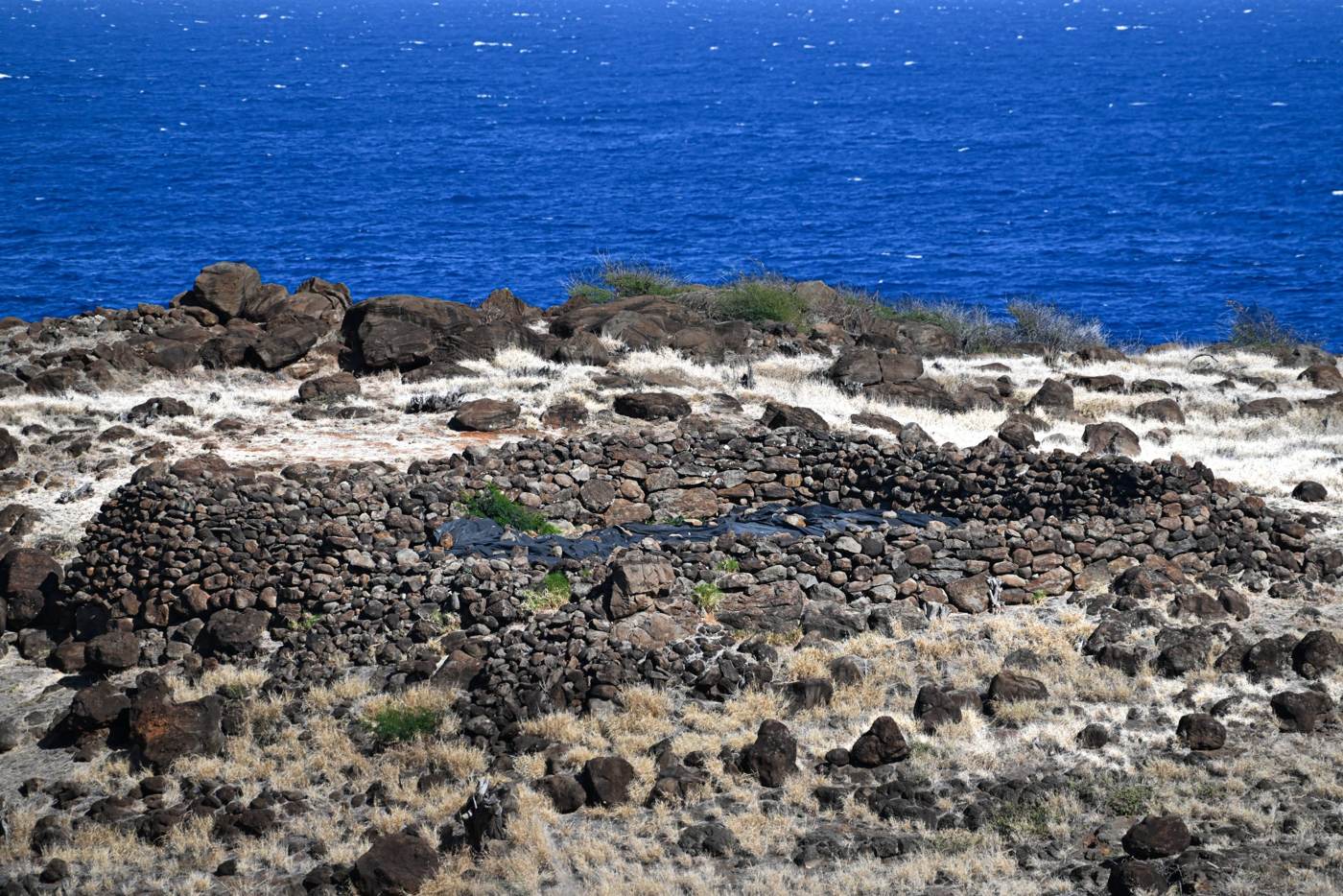 A stone structure surrounded by dry grass and rocks stands on a coastal landscape with the blue ocean in the background.