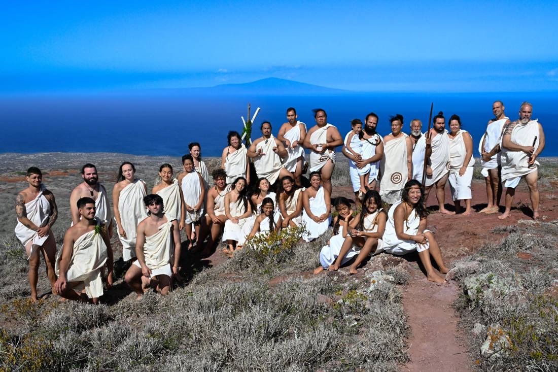 A group of people dressed in traditional attire pose on a rocky hillside with the ocean and a distant island visible under a clear blue sky.