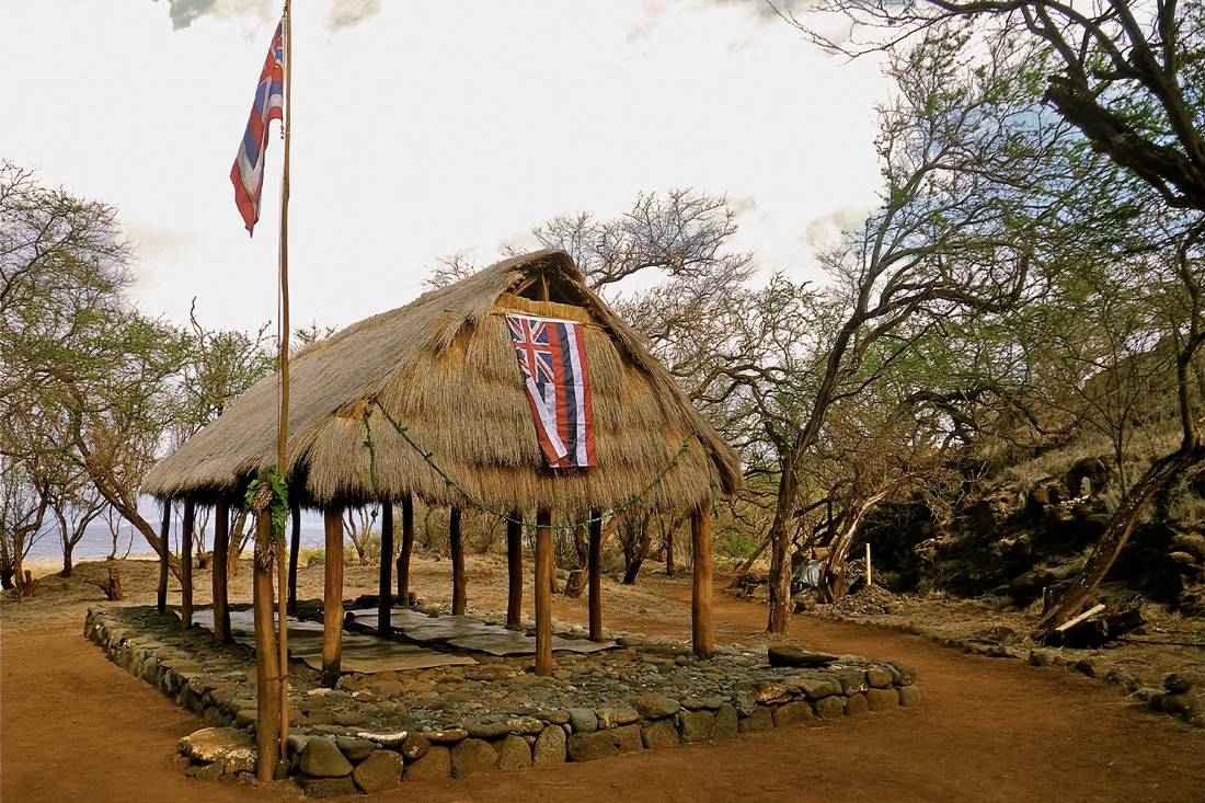 A traditional Hawaiian thatched hut stands on a stone foundation, adorned with the Hawaiian flag, surrounded by dry trees and earth.
