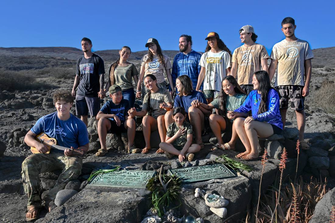 Thirteen people pose together outdoors on rocky terrain near two large plaques, some sitting and some standing, with one person playing a small stringed instrument.