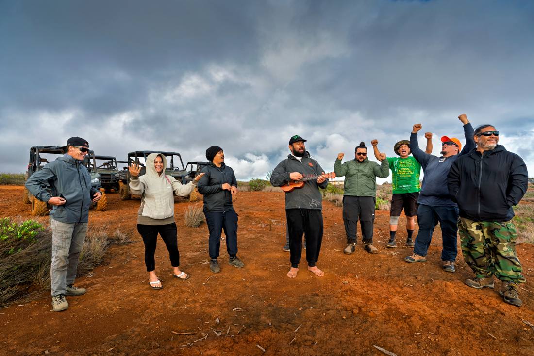 A group of people stand on a red dirt landscape with off-road vehicles in the background; one person plays a ukulele while others gesture and smile.